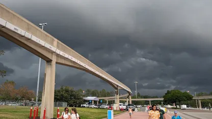 Elevated concrete transit track curves across a park beneath dark storm clouds, with pedestrians and vehicles below.