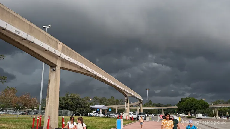 Elevated concrete transit track curves across a park beneath dark storm clouds, with pedestrians and vehicles below.