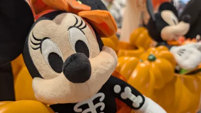 Minnie Mouse plush toy in an orange hat and black outfit, with autumn-themed decorations in the background.