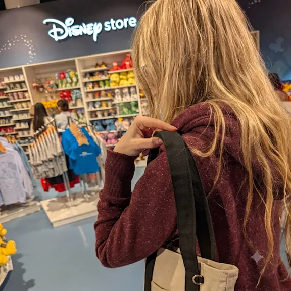 Woman browsing clothing in a shop, viewed from behind, with shelves of folded garments and mannequins in the background.