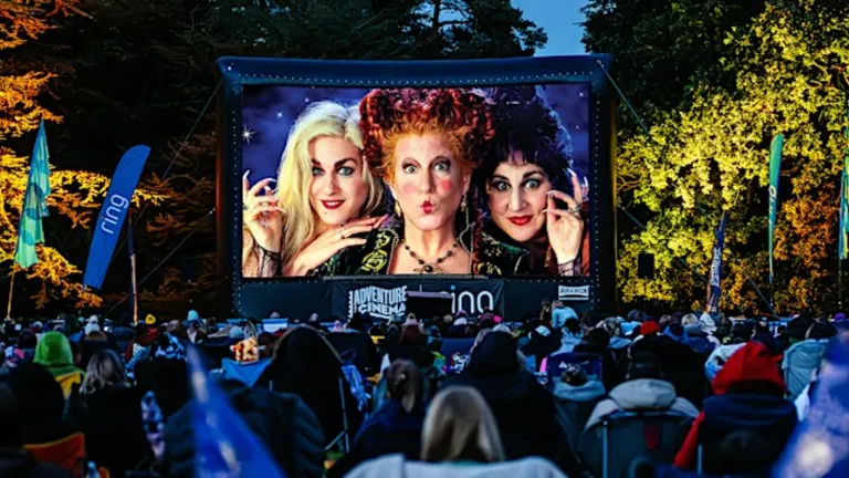 Outdoor film screening showing three witches on a large screen with an audience watching.