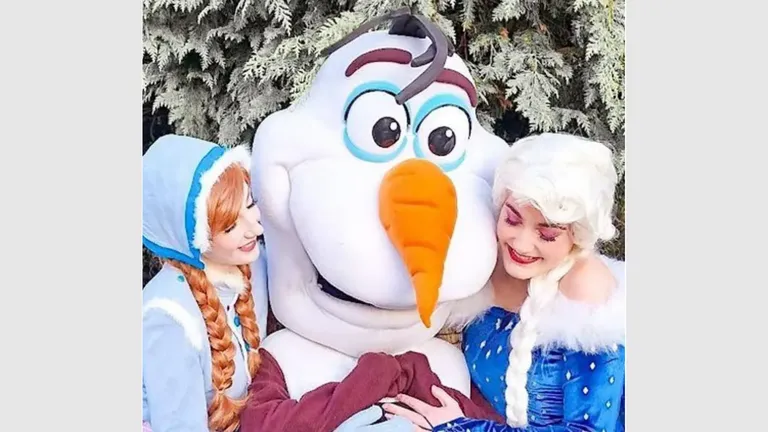 Elsa and Anna pose with Olaf, a snowman character, in front of frosted trees.