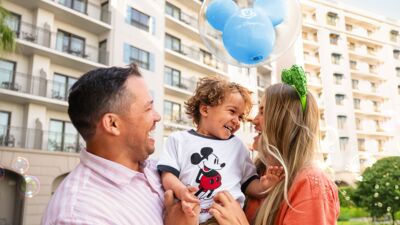 Family of three holding a child with Mickey Mouse balloons in front of a building