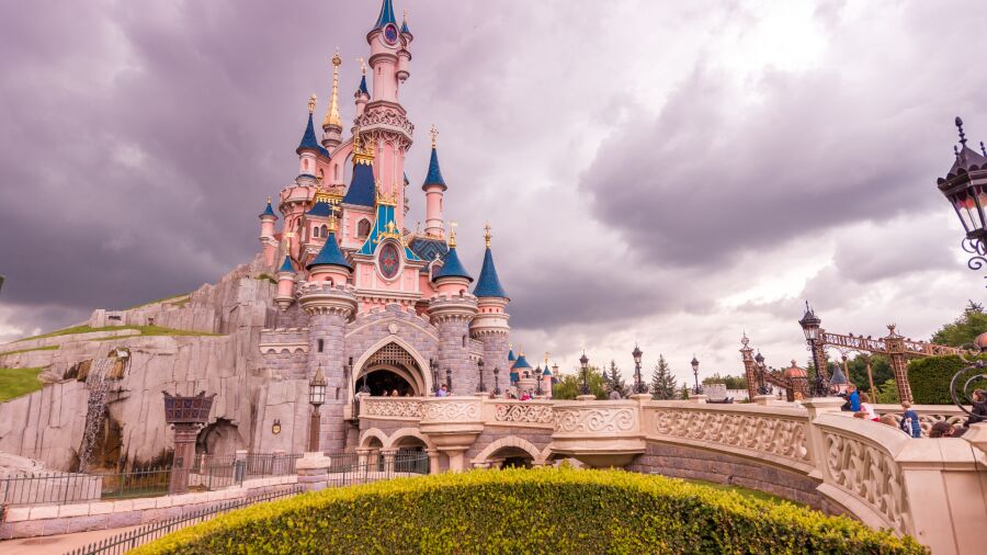 Disneyland Paris castle with blue and pink turrets, a bridge, and a cloudy sky