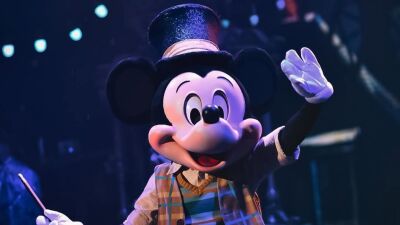 Mickey Mouse is waving and smiling while holding a wand and wearing a hat